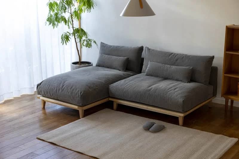 A sectional sofa with a chaise lounge component. The sofa features deep, plush seating and back cushions upholstered in a dark gray fabric. It is supported by a minimalist, light-colored wooden frame with tapered legs. — 3D model