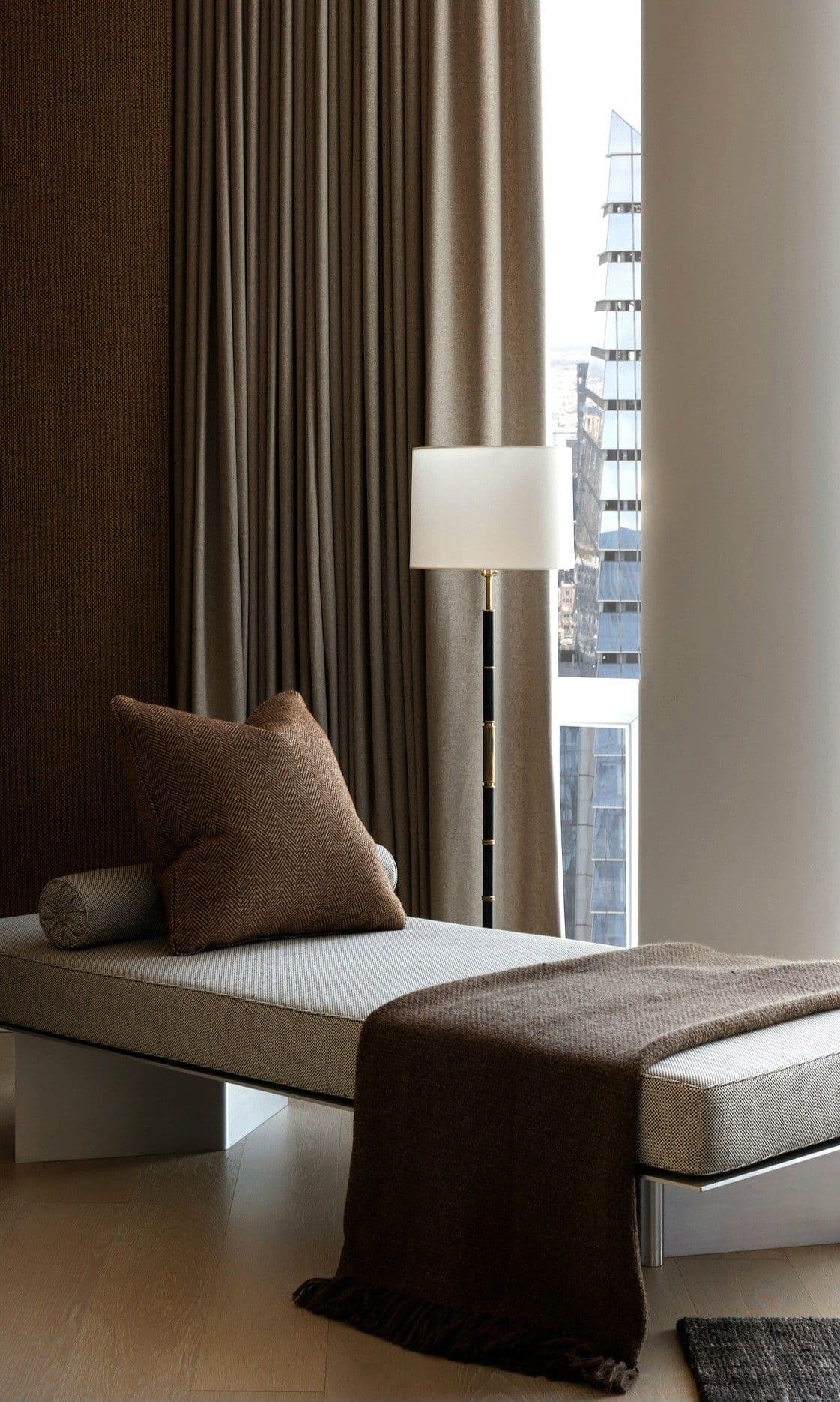 A modern daybed featuring a long, rectangular upholstered surface in a light neutral fabric, supported by a minimalist metal base. It is accessorized with a cylindrical bolster cushion, a square throw pillow, and a fringed throw blanket, all in a contrasting brown textile. — 3D model