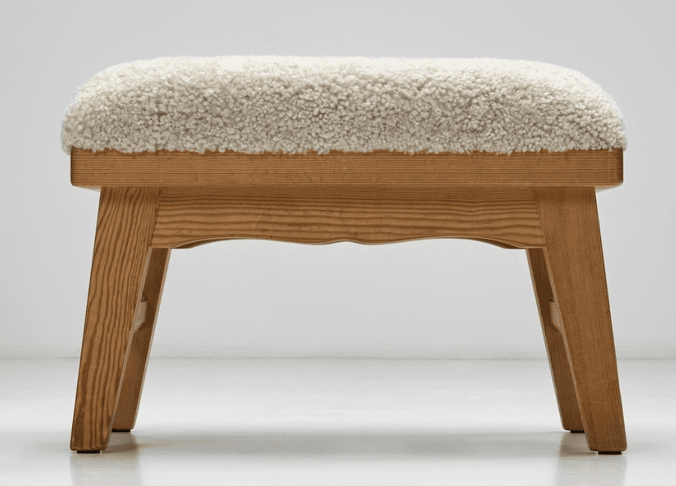 A rectangular stool featuring a thick, plush, cream-colored sheepskin upholstery top over a natural light-toned wooden frame. The frame has visible grain, sturdy splayed legs, and a subtly scalloped apron detail on the long sides. — 3D model