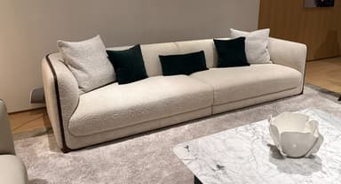A long, contemporary sofa featuring a distinctive sloping backrest upholstered in a light, textured cream fabric. The edges of the sofa, including the unique angled back, are accentuated with dark brown piping. It includes several throw pillows, some matching the main upholstery and others in a contrasting dark green velvet. The sofa rests on a recessed plinth base with a dark wood finish. — 3D model