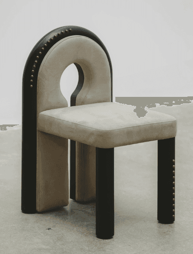 A contemporary chair with a distinctive U-shaped backrest that forms a cutout. The seat and the curved portion of the backrest are upholstered in a light beige, suede-like fabric. The frame is composed of dark, possibly black or dark wood, elements. The outer curved frame of the backrest and the vertical supports of the legs are adorned with a series of small, brass or gold-colored studs. The legs are thick and rectangular, with a dark finish. The chair's design is modern and somewhat sculptural, featuring a negative space in the upper backrest. — 3D model