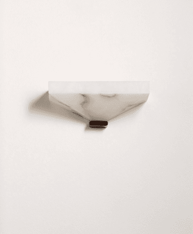 A contemporary wall sconce crafted from translucent alabaster, featuring a faceted, geometric form. The body is an inverted, truncated pyramid shape with a flat top, designed to cast light primarily upwards or downwards, depending on orientation. It exhibits natural veining inherent to alabaster, contributing to its organic yet refined aesthetic. A small, dark, rectangular base element is visible at the bottom, likely serving as a mounting point or decorative accent. The overall design suggests a sophisticated and minimalist lighting solution. — 3D model
