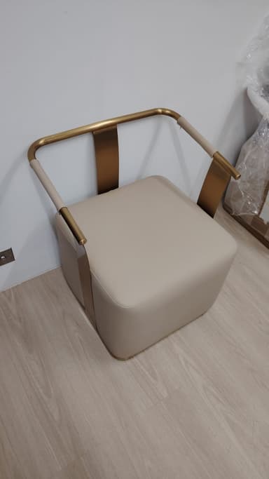 A modern armchair featuring a square, thick cushioned seat upholstered in a light beige material, supported by a minimalist brass-toned tubular metal frame that seamlessly forms the backrest and armrests. The design is sleek and contemporary. — 3D model