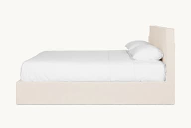 A bed frame with a padded headboard and platform base, covered in a neutral-toned fabric. The headboard has a distinctive silhouette with curved upper corners. The bed is shown with white linens and two pillows. — 3D model