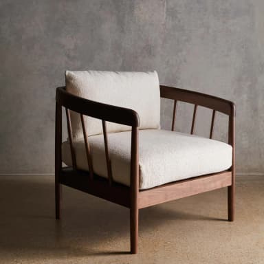 A side profile view of a wooden armchair featuring a curved backrest and slatted sides. The seat and a single throw pillow are upholstered in a textured, light-colored fabric. — 3D model
