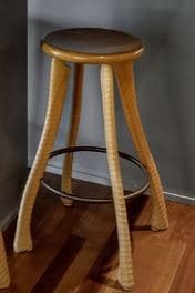 A tall, contemporary bar stool featuring a round, padded dark brown seat. Its three splayed legs are constructed from wood and meticulously wrapped in a light-colored woven natural fiber, likely rattan or cane, creating a textured surface. A black circular metal footrest connects the legs, providing stability and support. — 3D model