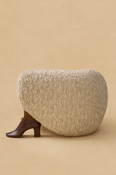 An armless lounge chair featuring a fully upholstered body in a cream-colored, textured bouclé fabric, supported by four dark, organically shaped wooden legs. The chair has a low profile and a rounded, inviting silhouette. — 3D model