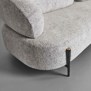 A close-up view of a segment of a modern sofa, featuring rounded, organic lines and textured upholstery. The sofa has a plush appearance, with a low profile and substantial cushioning. A single, distinctly designed metal leg is visible, supporting the rounded front base of the sofa. The leg has a dark, textured finish with a cap that appears to be a different material, possibly metal or wood with a metallic finish. — 3D model