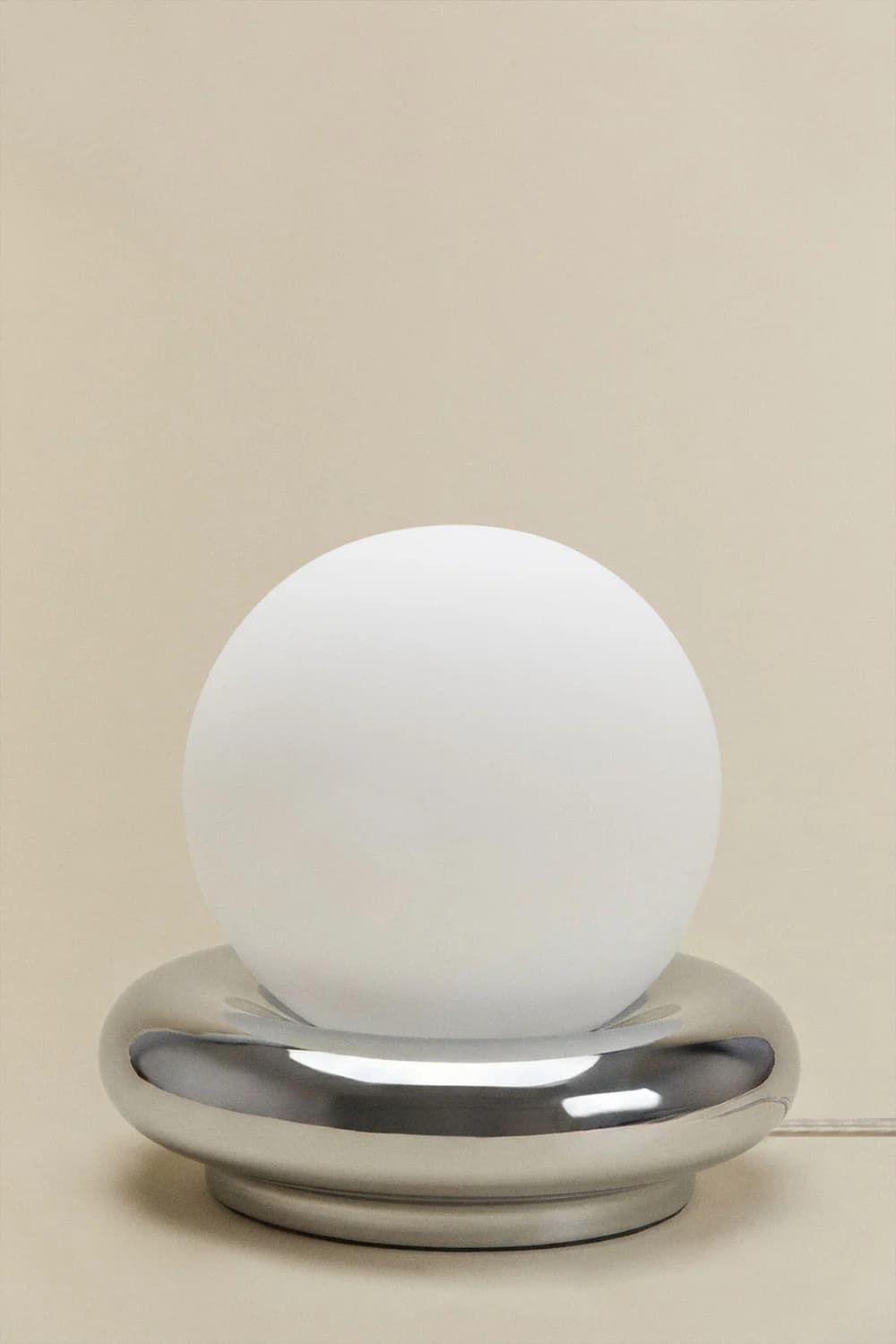 A modern table lamp featuring a spherical frosted white glass diffuser resting within a polished chrome base with a rounded, toroidal shape. The minimalist design emphasizes geometric forms and a sleek, reflective finish on the base, providing ambient light through the globe. — 3D model