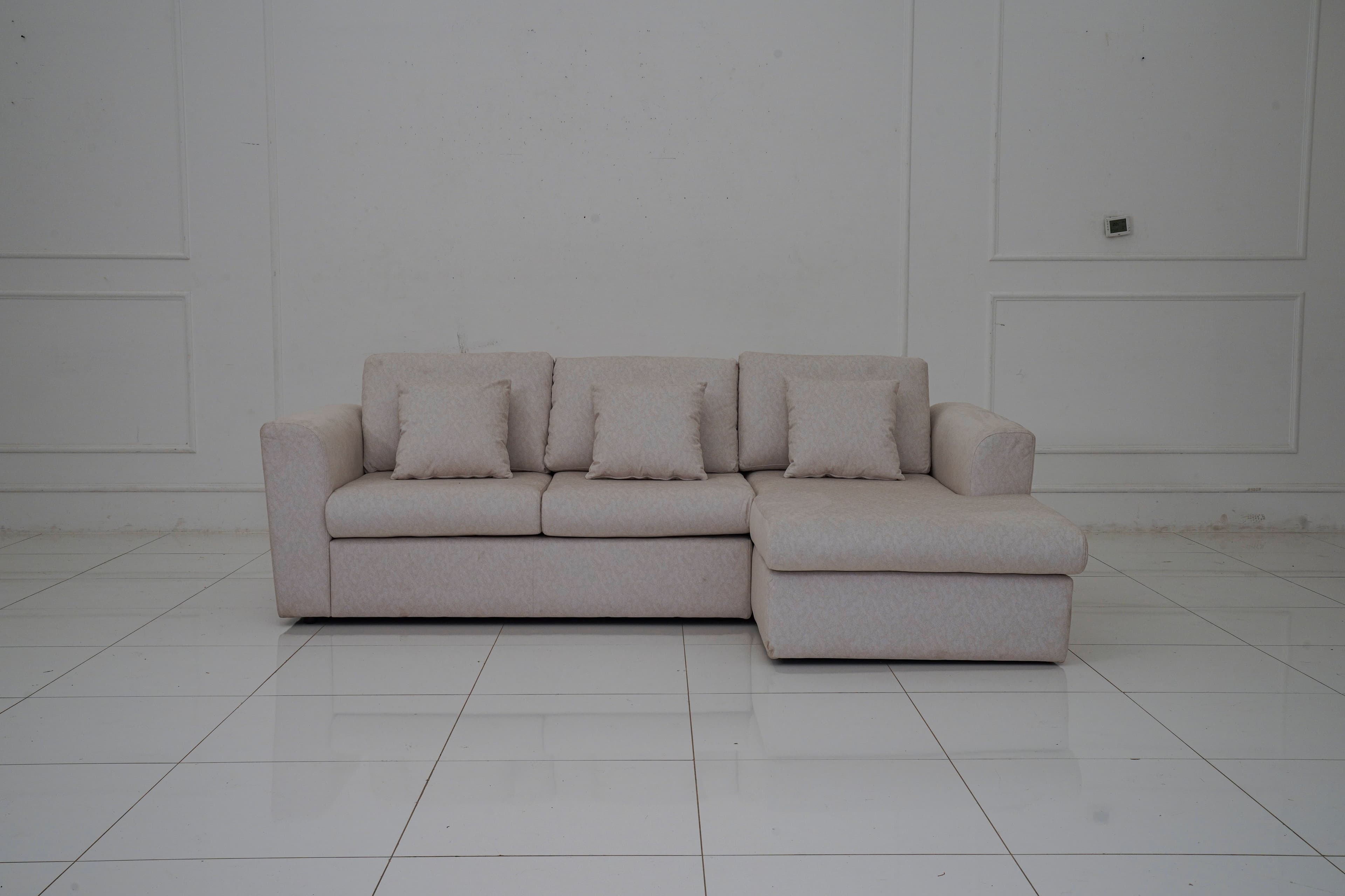 A sectional sofa with a chaise lounge extension, upholstered in a light-colored fabric with a subtle pattern. The sofa features three seat cushions and a chaise on the right side. It includes four matching throw pillows. The sofa has a modern, minimalist design with clean lines and low-profile arms and back. — 3D model