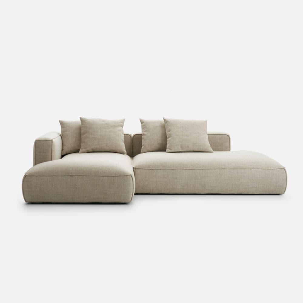 A contemporary modular sectional sofa featuring a low-profile design and upholstered in a light beige textured fabric. It consists of two main pieces: a corner section with an armrest and a chaise lounge extension, creating an L-shaped configuration. The sofa is characterized by its soft, rounded edges, generous proportions, and deep seating. It comes with several loose back cushions, adding to its plush appearance and comfort. — 3D model