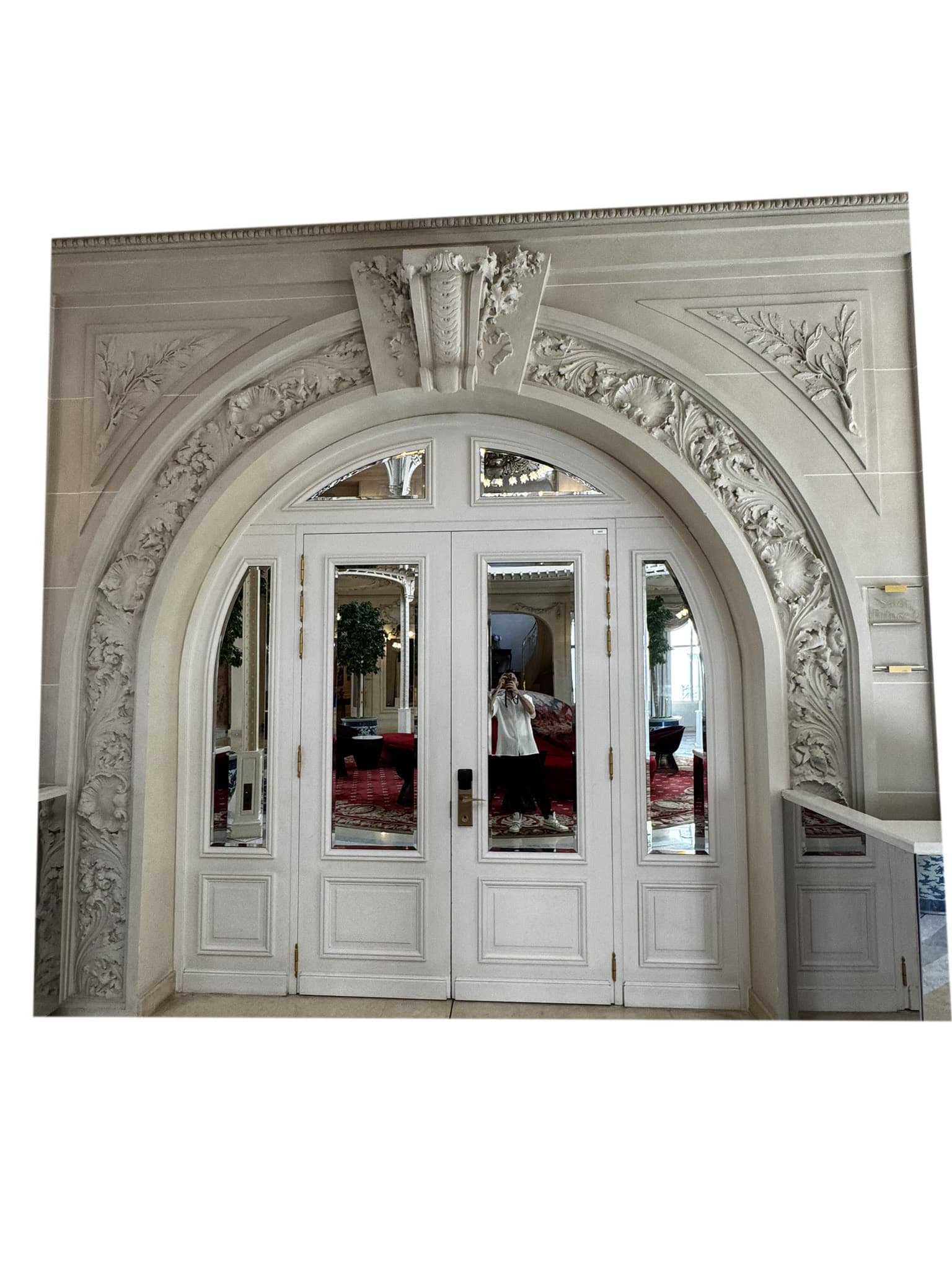 A large, ornate, double-door entryway framed by an elaborate arched stone surround. The doors are off-white or light beige, made of wood or a wood-like material, with glass panels and brass hardware. The stone surround features high-relief carvings of floral and foliate patterns in a style suggestive of classicism or neoclassicism. The overall condition appears to be good, with the materials showing minimal visible wear. — 3D model