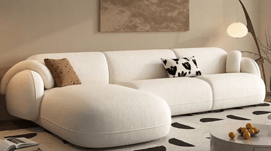 A sectional sofa with a cream-colored fabric upholstery. It features rounded arms and a low profile. The sofa is configured in an L-shape, with one section appearing to be a chaise lounge. Two decorative throw pillows are placed on the sofa: one with a brown textured pattern and the other with a black and white abstract design. — 3D model