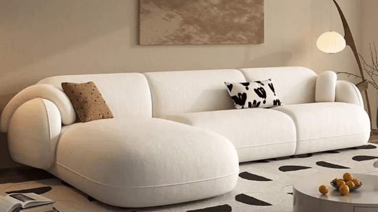 A sectional sofa with a cream-colored fabric upholstery. It features rounded arms and a low profile. The sofa is configured in an L-shape, with one section appearing to be a chaise lounge. Two decorative throw pillows are placed on the sofa: one with a brown textured pattern and the other with a black and white abstract design. — 3D model