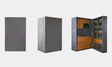 A freestanding, rectangular cabinet with a dark gray, textured concrete-look exterior featuring an embossed geometric pattern. The cabinet opens to reveal a fully integrated compact kitchen and bar area, complete with wooden cabinetry, a sink, a stovetop, a microwave, and shelving designed for glassware and bottles. — 3D model