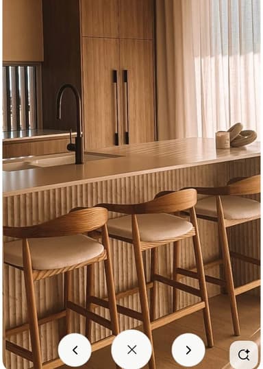 A modern bar stool with a light wood frame and a beige upholstered seat, featuring a curved backrest that doubles as armrests. The legs are slightly splayed with cross-bracing for stability. — 3D model