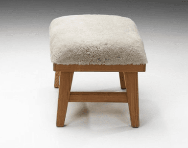A rectangular stool featuring a thick, plush, cream-colored sheepskin upholstery top over a natural light-toned wooden frame. The frame has visible grain, sturdy splayed legs, and a subtly scalloped apron detail on the long sides. — 3D model