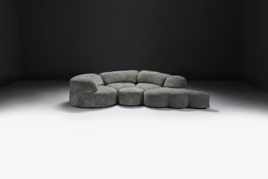 A large, multi-part sectional sofa featuring soft, organic, rounded forms, upholstered in a textured grey fabric. The sofa comprises several modules including corner pieces, armless central sections, and an end module that functions as an extended footrest or chaise. The overall design emphasizes comfort and a flowing, contemporary aesthetic. — 3D model