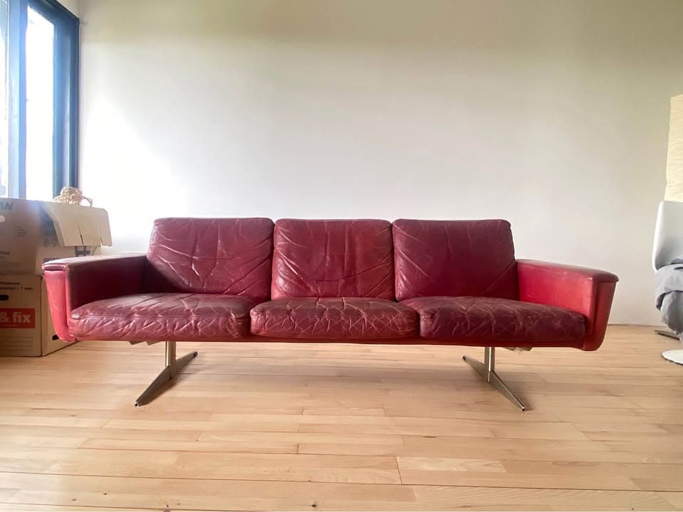 A three-seater sofa with a low profile and square arms. The sofa is upholstered in a red, textured material, likely leather, and features distinct cushion divisions. It is supported by slender, angled metal legs. — 3D model