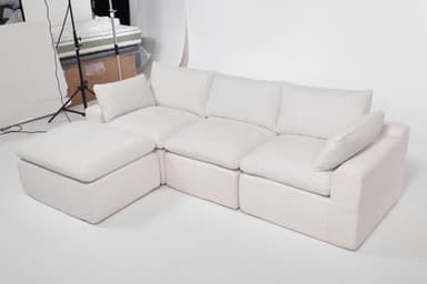 A modular sectional sofa with an attached ottoman. The sofa features deep, plush cushions and a low profile. It is upholstered in a textured white fabric. The configuration shown includes a corner section, two seat cushions, and an ottoman. The design is contemporary and emphasizes comfort. — 3D model