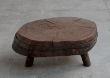 A rustic and primitive low table, likely serving as a coffee table or accent table, crafted from a thick, irregularly shaped slab of wood, resembling a cross-section of a tree stump. The top surface displays prominent wood grain and natural radial cracks, indicating its organic origin and age. A distressed metal band with visible rivets encircles the perimeter, securing the wood and contributing to its rugged aesthetic. The table stands on three short, sturdy, tapered wooden legs. — 3D model
