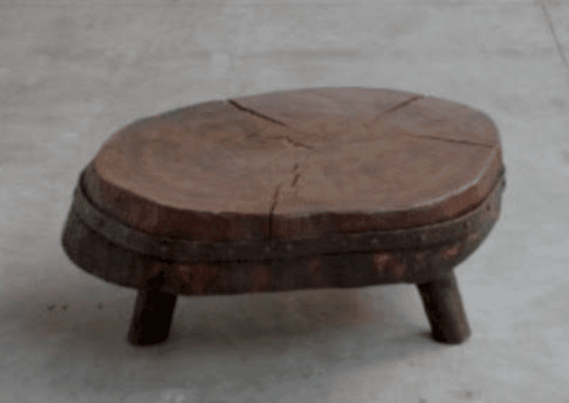 A rustic and primitive low table, likely serving as a coffee table or accent table, crafted from a thick, irregularly shaped slab of wood, resembling a cross-section of a tree stump. The top surface displays prominent wood grain and natural radial cracks, indicating its organic origin and age. A distressed metal band with visible rivets encircles the perimeter, securing the wood and contributing to its rugged aesthetic. The table stands on three short, sturdy, tapered wooden legs. — 3D model