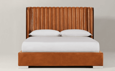 A contemporary platform bed featuring a tall, curved headboard with vertical fluted cognac leather upholstery, flanked by dark wood side panels. The bed frame is also upholstered in matching cognac leather, resting on polished metal block feet. — 3D model