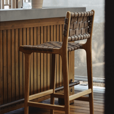 A bar stool with a light-colored wooden frame and woven leather straps for the seat and backrest. The wooden frame has a smooth, curved design, and the legs are connected by stretchers for stability. The leather straps are interlaced to form a textured surface for seating and back support. A patterned rug is visible beneath the stool. — 3D model