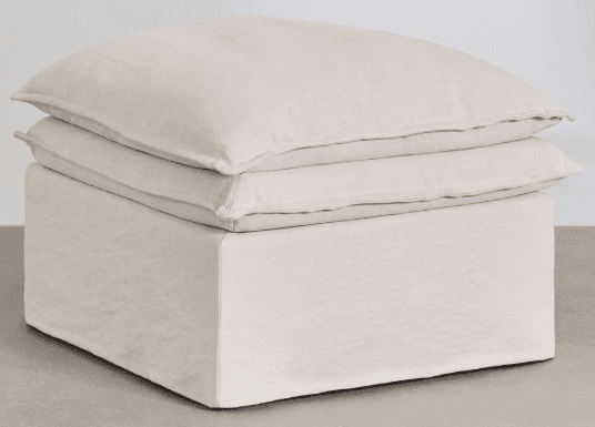 A light beige, square, slipcovered ottoman featuring a boxy base and two stacked, plush cushions on top, presenting a soft and inviting appearance. — 3D model