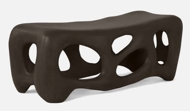 A modern, abstract bench with a dark brown, matte finish. The design features organic, irregular openings throughout its structure, creating a porous or skeletal appearance. Its form is elongated and somewhat fluid, with rounded edges and a low profile, resting on integrated leg-like supports that are part of the overall molded form. — 3D model