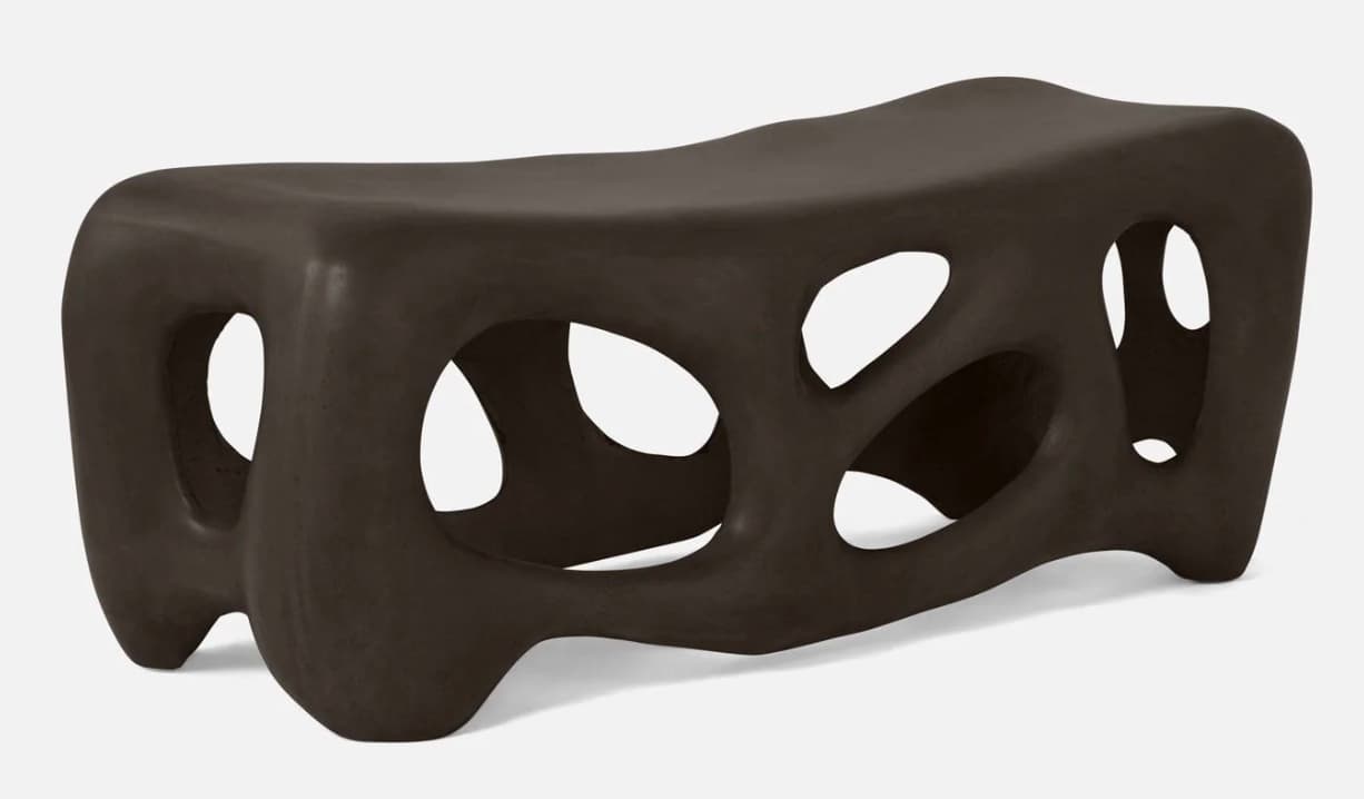 A modern, abstract bench with a dark brown, matte finish. The design features organic, irregular openings throughout its structure, creating a porous or skeletal appearance. Its form is elongated and somewhat fluid, with rounded edges and a low profile, resting on integrated leg-like supports that are part of the overall molded form. — 3D model