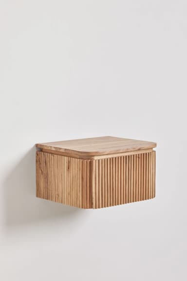 A rectangular, wall-mounted wooden nightstand featuring a natural wood finish. The front and sides of the unit are adorned with a vertical reeded or fluted texture, providing a tactile and decorative element. The top surface is smooth with gently rounded edges, slightly overhanging the reeded body. A subtle seam on the front suggests a drawer or hinged door for internal storage. — 3D model