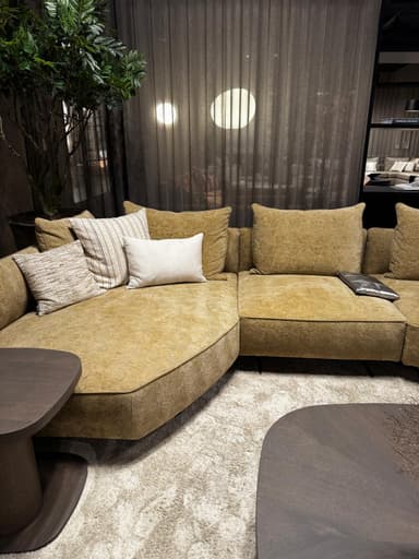 A sectional sofa with a textured, mustard-yellow fabric upholstery. The sofa features a low profile and deep seating. It is adorned with several throw pillows, including one in a neutral tone and others with subtle striped and textured patterns. The base of the sofa has a distinctive geometric pattern. Two dark wood coffee tables with rounded edges are positioned in front of the sofa, and a plush, light-colored rug covers the floor. — 3D model