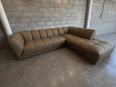 An L-shaped sectional sofa featuring a right-hand facing chaise, upholstered in a smooth, light brown synthetic leather. The design includes distinct biscuit tufting across all seat and back cushions, creating a plush, segmented appearance, and is characterized by a low-profile silhouette with wide, integrated armrests. — 3D model