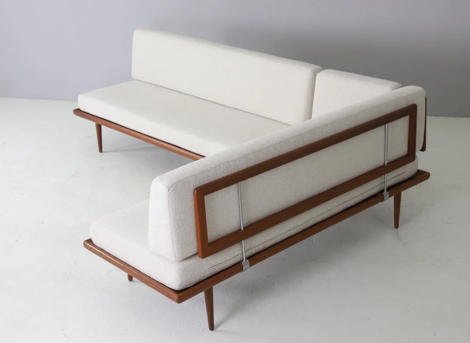 An L-shaped sectional sofa featuring an exposed dark-stained wooden frame, likely teak, supporting thick, light-colored upholstered cushions. The sofa consists of two distinct segments forming a right-angle configuration. The backrests are also upholstered and appear to be slightly reclined, secured within the wooden frame with visible metal supports. The frame rests on slender, tapered wooden legs. — 3D model