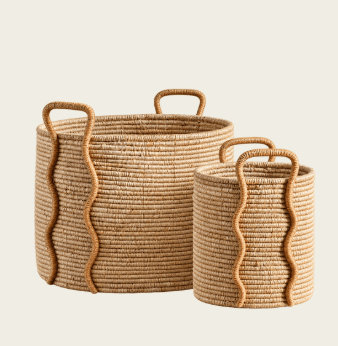 A cylindrical storage basket crafted from tightly coiled natural fibers, likely jute or seagrass, presenting a natural light brown color. It features two integrated loop handles at the rim for portability and two decorative wavy vertical rope accents on the exterior sides. The basket has an open-top design and a sturdy base, suitable for various storage needs. — 3D model