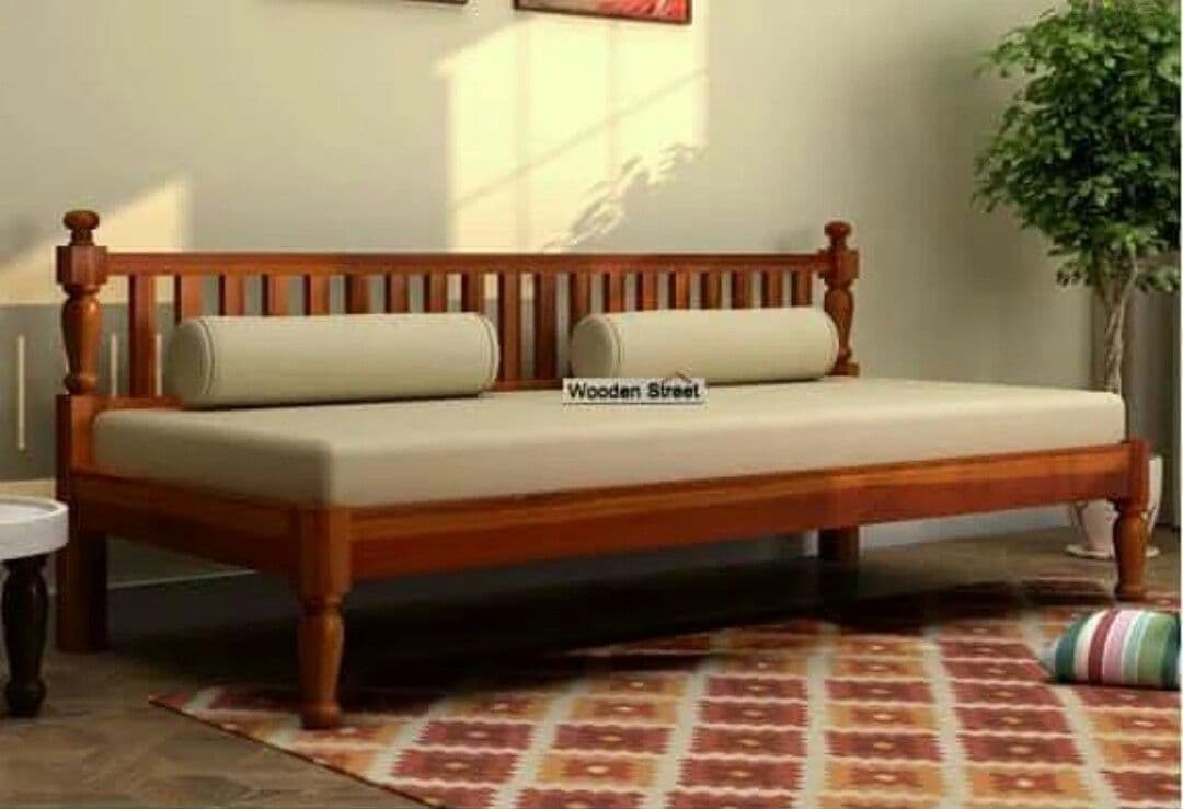 A traditional wooden daybed featuring a slatted backrest and turned legs, presented with a beige mattress and two matching bolster pillows. The frame is constructed from a reddish-brown wood, indicative of teak or a similar hardwood, with decorative finials on the backrest posts. The overall design emphasizes functionality and classic aesthetics. — 3D model