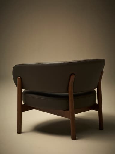 A low-profile armchair with a dark brown leather upholstered seat and curved backrest. The frame and legs are constructed from dark wood, exhibiting a minimalist design. The seat and back appear to be separate components supported by the wooden frame. — 3D model