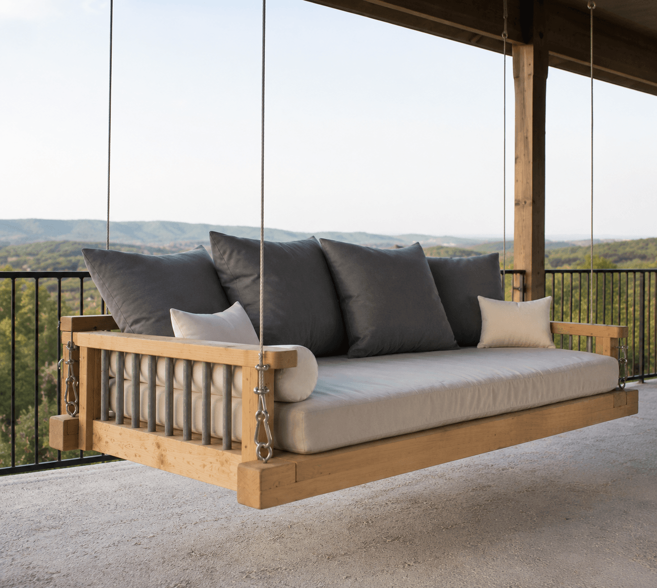 A modern, light-toned wooden suspended daybed featuring a thick off-white mattress cushion, multiple dark grey throw pillows, and white bolster cushions, all suspended by four metal chains. — 3D model