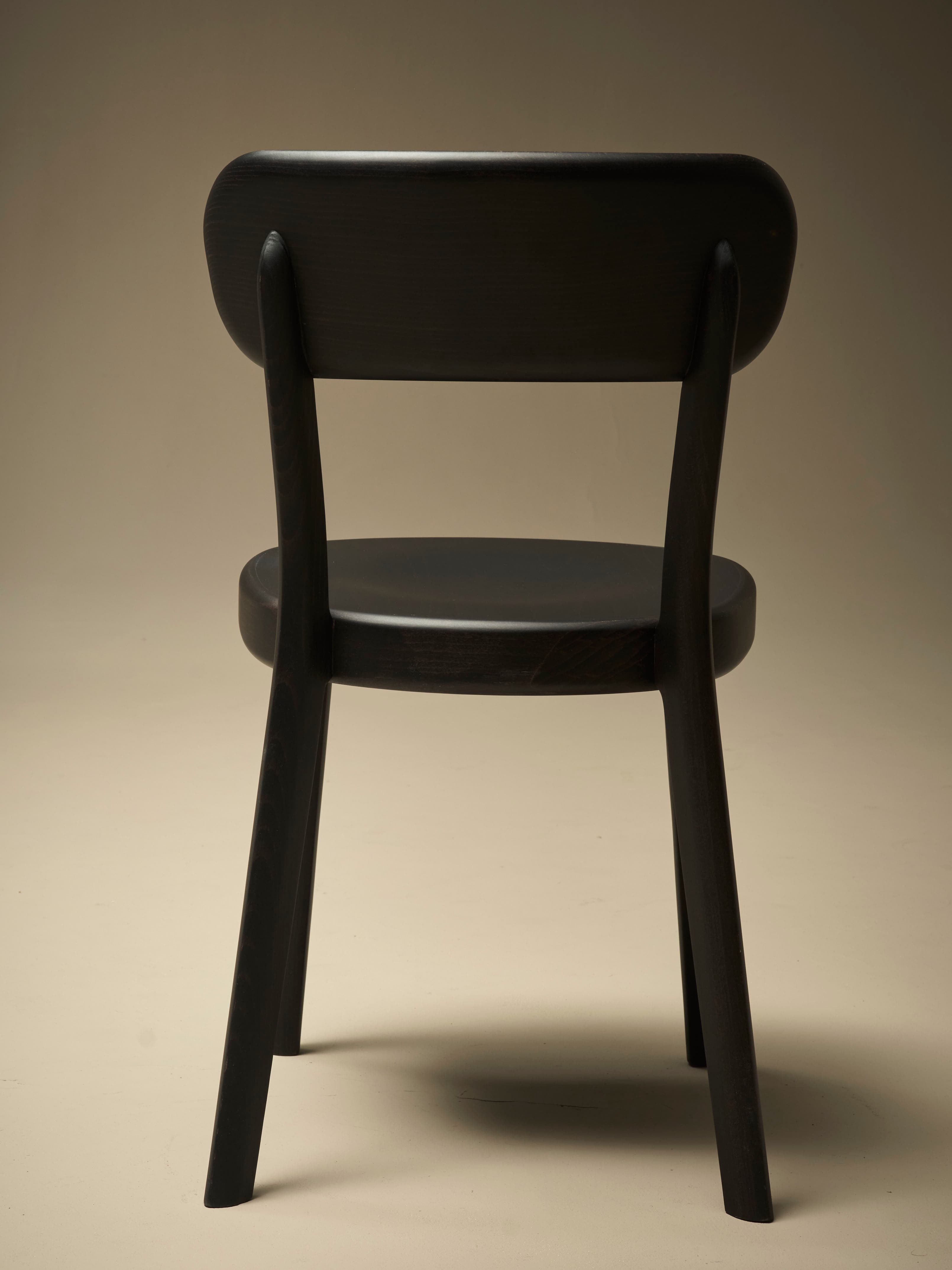 A dark-colored wooden dining chair viewed from the back. The chair features a gently curved backrest supported by two upright posts that extend from the seat. The seat itself is also curved and appears to be solid. The legs are slightly tapered and angled outwards. The overall construction suggests a modern or contemporary design with clean lines and a minimalist aesthetic. — 3D model