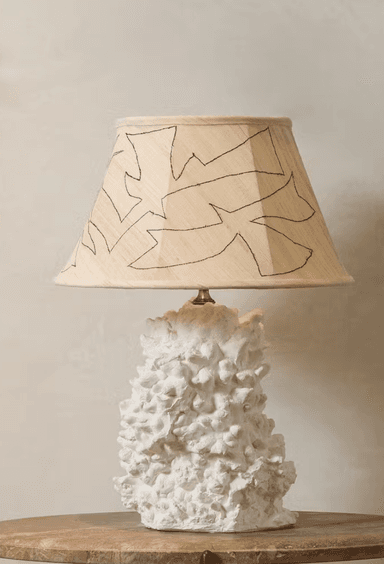 A contemporary table lamp featuring a sculptural, highly textured white ceramic or plaster base that resembles organic forms like coral or abstract rock formations. It is topped with a conical fabric lampshade in a neutral beige or cream color, adorned with a black outline illustration depicting abstract leaves or organic shapes. The overall design emphasizes natural textures and artistic detailing. — 3D model