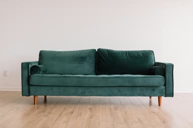 A modern three-seater sofa upholstered in a rich emerald green velvet fabric. It features a button-tufted seat cushion, two large back cushions, and narrow square armrests, each accompanied by a matching cylindrical bolster cushion. The sofa stands on four tapered, light-toned wooden legs. — 3D model
