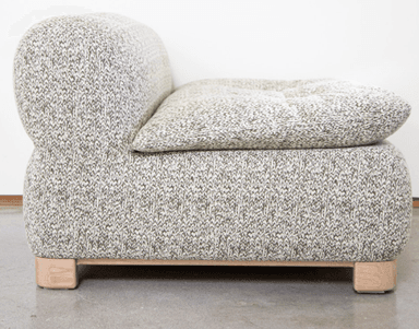 A contemporary armchair with a distinctive rounded silhouette. The piece is upholstered in a textured knit fabric with a marled pattern of light grey and white. It features a low profile, a generously padded seat cushion, and a softly curved backrest. The armchair is supported by a minimalist wooden base with four block-like feet. — 3D model