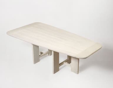 A rectangular dining table crafted from light-colored wood, featuring a slatted top with gently rounded corners. The table is supported by two distinct trestle-style bases, each composed of two vertical elements braced by a horizontal stretcher near the floor. The wood exhibits a bleached or limed finish, emphasizing its grain and contributing to a minimalist aesthetic. — 3D model