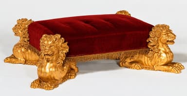 A rectangular ottoman featuring a deeply button-tufted red velvet upholstered top, supported by four intricately carved and gilded lion figures serving as legs. The upholstery is accented with a delicate gold fringe trim along its lower edge. — 3D model