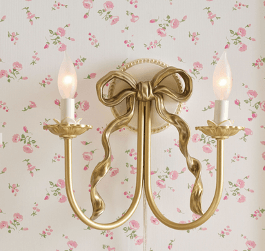 A decorative wall sconce designed to resemble a candle holder with a bow motif. It features two candle-shaped light bulbs emitting a warm glow. The structure is supported by curved arms extending from a central base adorned with a sculpted ribbon or bow. — 3D model