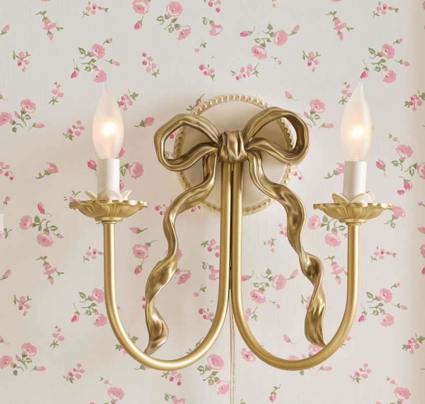 A decorative wall sconce designed to resemble a candle holder with a bow motif. It features two candle-shaped light bulbs emitting a warm glow. The structure is supported by curved arms extending from a central base adorned with a sculpted ribbon or bow. — 3D model