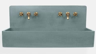 A wall-mounted, rectangular trough sink made of blue-gray concrete, featuring an integrated backsplash. Two sets of wall-mounted brass faucets with cross handles are installed above the basin. The design is minimalist and industrial. — 3D model