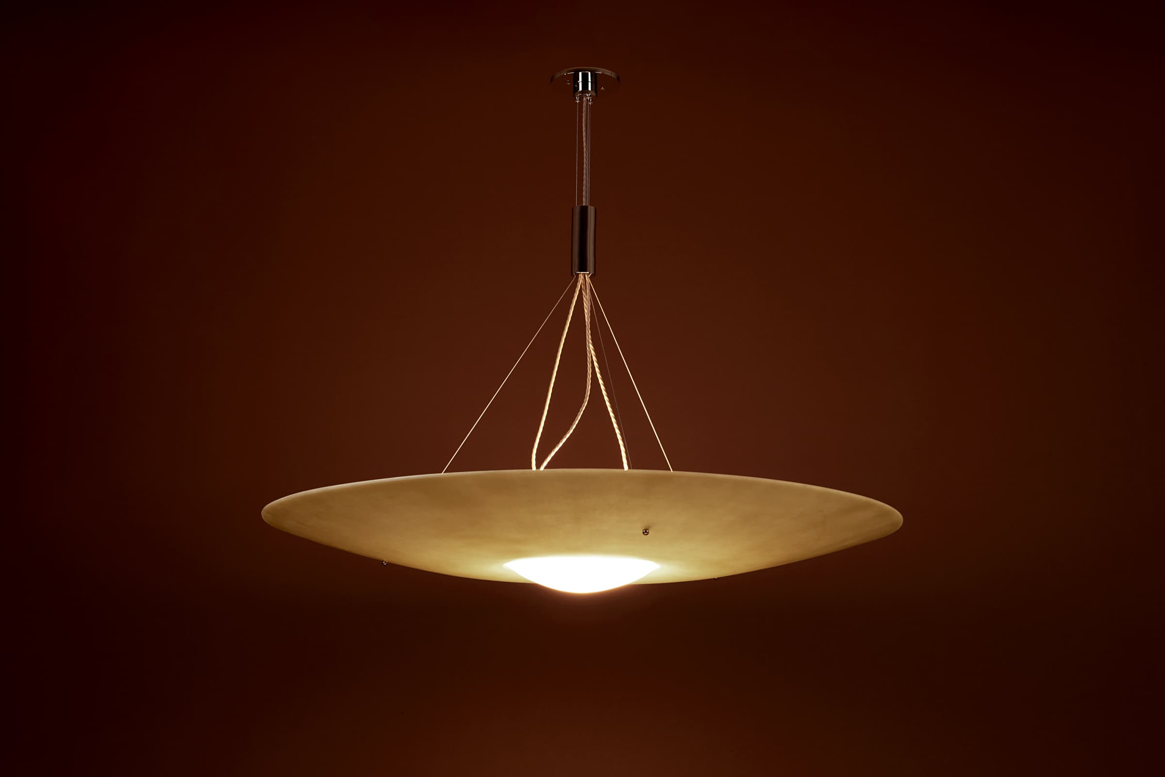 A contemporary chandelier featuring a wide, flattened disc-shaped shade in an off-white or cream color. The shade has a slightly upward-curving lip and an integrated light source at its center, which emits a warm glow. It is suspended from the ceiling by a thin cable and a metallic rod/canopy assembly, with multiple finer cords attaching the shade to the central support. — 3D model