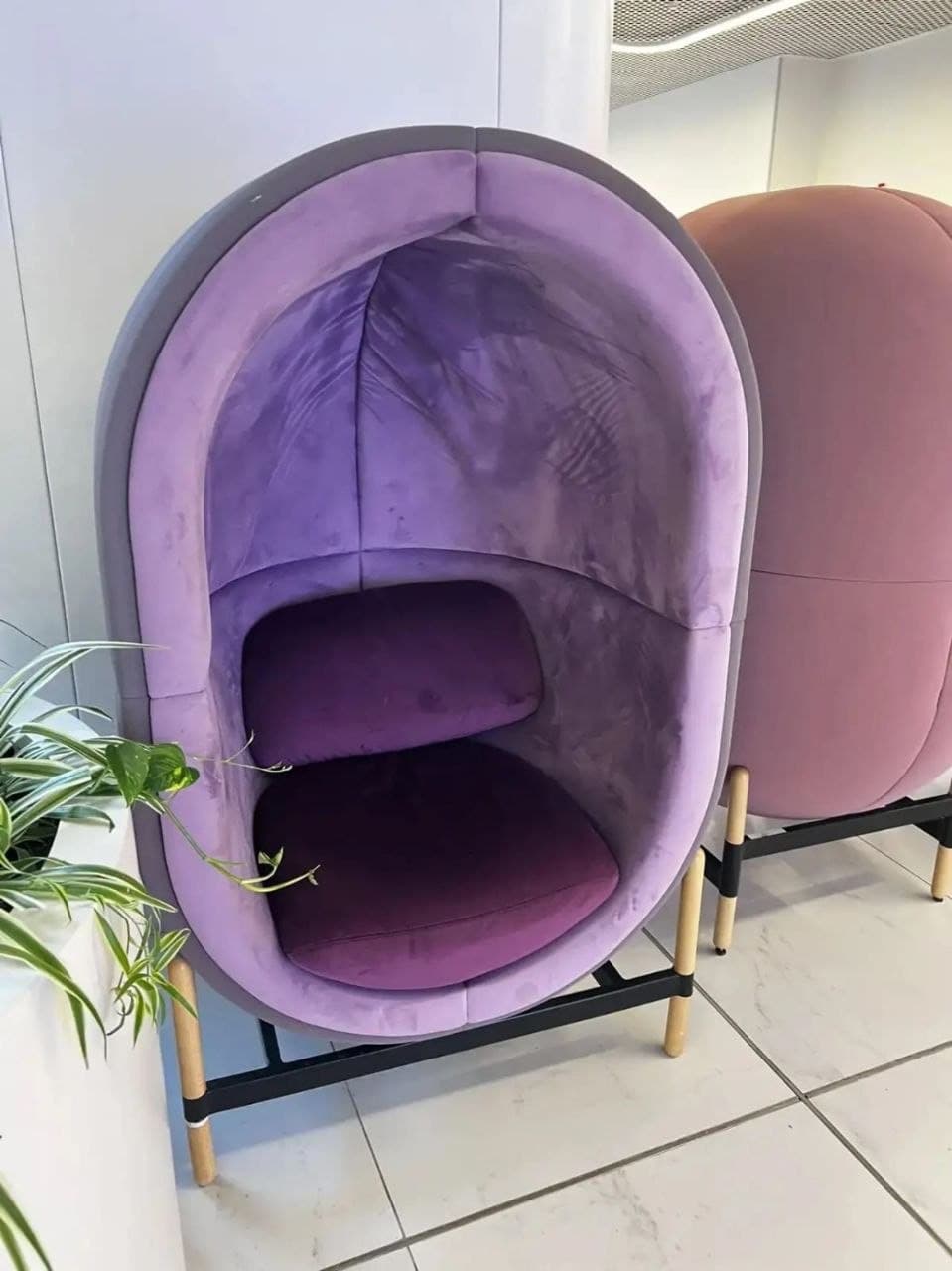 An upholstered pod privacy lounge chair with an egg-shaped exterior in grey and a lavender-purple interior. It stands on splayed wooden legs connected by black metal crossbars. — 3D model